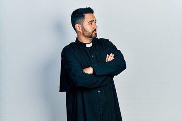 Handsome hispanic man with beard wearing catholic priest robe looking to the side with arms crossed...