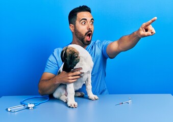 Handsome hispanic veterinary man with beard checking dog health pointing with finger surprised...