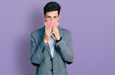Young hispanic business man covering face with cards relaxed with serious expression on face....