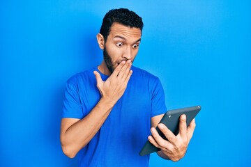 Hispanic man with beard using touchpad device covering mouth with hand, shocked and afraid for...