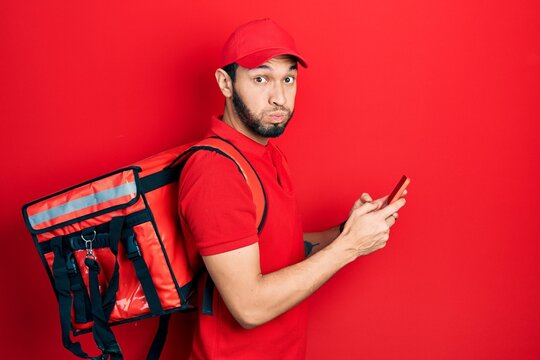 Hispanic man with beard wearing delivery package backpack using smartphone puffing cheeks with funny face. mouth inflated with air, catching air.