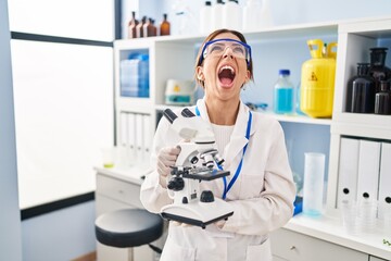 Young brunette woman working at scientist laboratory with microscope angry and mad screaming frustrated and furious, shouting with anger looking up.