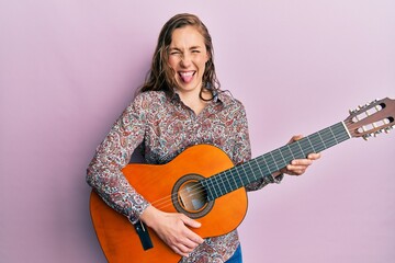 Young blonde woman playing classical guitar sticking tongue out happy with funny expression.