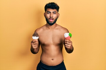 Fototapeta premium Arab man with beard wearing swimwear eating two ice cream cones afraid and shocked with surprise and amazed expression, fear and excited face.