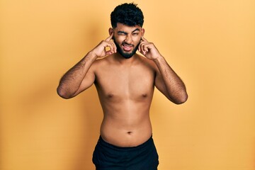 Fototapeta premium Arab man with beard wearing swimwear shirtless covering ears with fingers with annoyed expression for the noise of loud music. deaf concept.