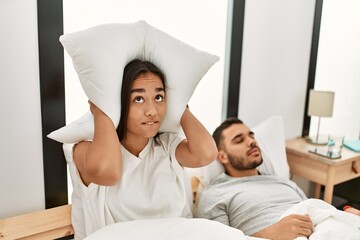 Woman covering ears with pillow irritated for snore lying in bed at home.