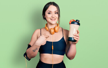 Young hispanic girl wearing sport clothes drinking a protein shake pointing finger to one self...