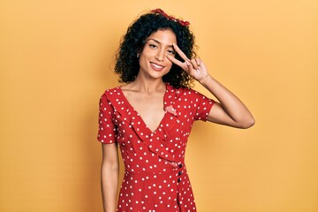 Young latin girl wearing summer dress doing peace symbol with fingers over face, smiling cheerful showing victory