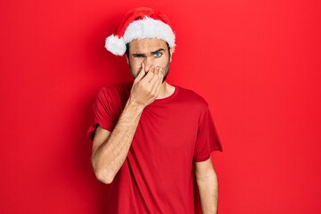 Young hispanic man wearing christmas hat smelling something stinky and disgusting, intolerable...
