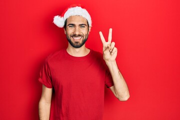Fototapeta premium Young hispanic man wearing christmas hat showing and pointing up with fingers number two while smiling confident and happy.