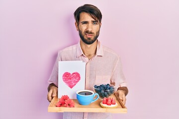 Young hispanic man holding tray with breakfast food and heart draw clueless and confused...