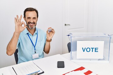 Middle age man with beard sitting by ballot holding i vote badge smiling positive doing ok sign with hand and fingers. successful expression.