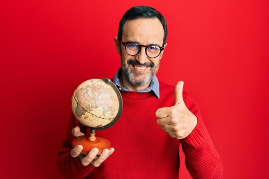 Middle age hispanic man holding vintage world ball smiling happy and positive, thumb up doing excellent and approval sign