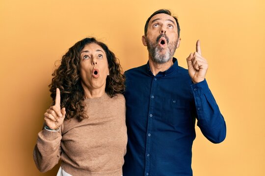 Middle age hispanic couple wearing casual clothes amazed and surprised looking up and pointing with fingers and raised arms.