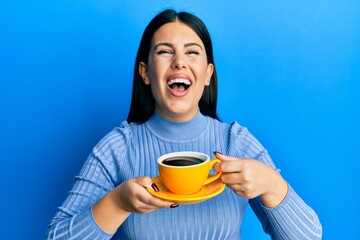 Beautiful brunette woman drinking a yellow cup of black coffee smiling and laughing hard out loud because funny crazy joke.