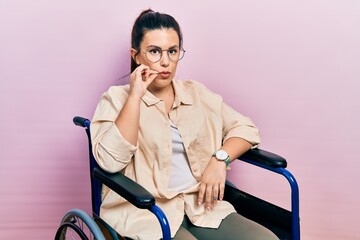Young hispanic woman sitting on wheelchair mouth and lips shut as zip with fingers. secret and...