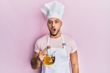 Young arab man wearing professional cook uniform holding olive oil can scared and amazed with open mouth for surprise, disbelief face