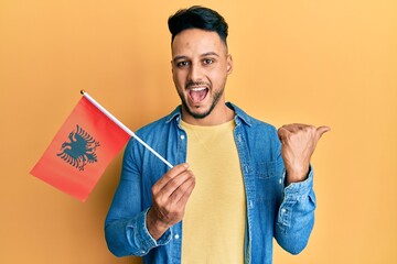 Young arab man holding albania flag pointing thumb up to the side smiling happy with open mouth - obrazy, fototapety, plakaty