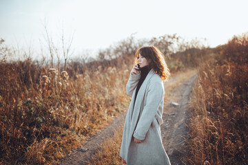 a red-haired curly-haired girl in a beige coat stands in a field among dried grass at sunset