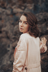 A girl in a pale pink raincoat and bra stands alone on a rock