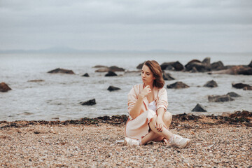 Fototapeta na wymiar A girl in a pale pink raincoat and a white T-shirt walks alone along the sandy beach