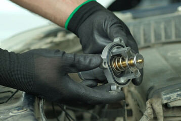 Repair and maintenance in a car service center. Spare parts. Thermostat for the engine. Close-up.  An auto mechanic holds a new thermostat in his hands against the background of the engine compartment