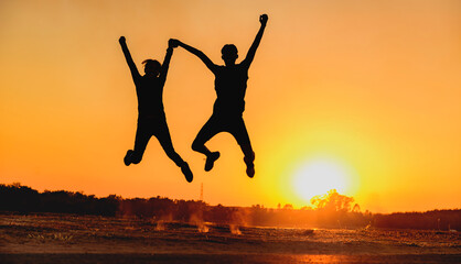 Male and female couple jumping happily at sunset with sun in the background, silhouette.