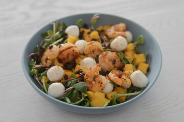 Salad with mango, mozzarella, shrimps and mixed microgreens in blue bowl on white oak table