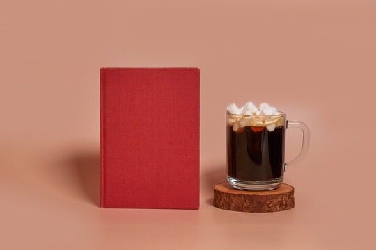 A glass cup of coffee with marshmallows stands on a wooden stand next to book