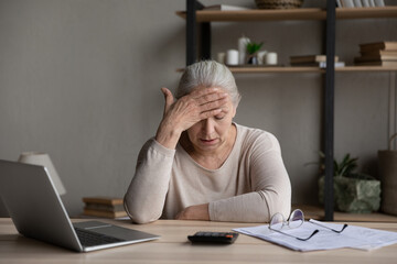 Worried upset older woman sit at table with laptop, counts expenses, manage budget, check bills for...