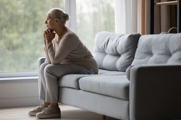 Fototapeta premium Side view upset older woman sit on sofa alone looking out window, thinking about problems, remembering moments of life, misses her children, feeling loneliness. Memories, grieving, solitude concept