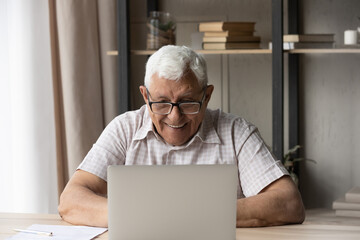 Happy smiling older man wear glasses sit at table at home read news staring on computer screen, use laptop learn information online, spend leisure using modern wireless tech and internet connection