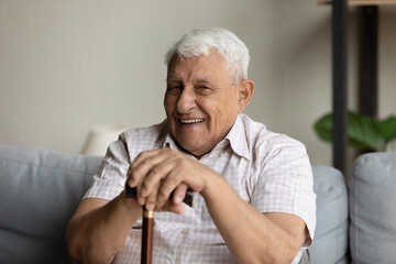 Physically disabled optimistic grandfather hold walking stick smile at camera, looks positive, rest alone on sofa at home. Healthcare, medical insurance for older citizen, recover after trauma concept