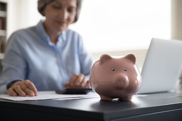 Classic piggy bank on desk, older woman sit at workplace with laptop use calculator counts monthly payments, on background. Taxes, incomes calculation, finances management, save money, savings concept