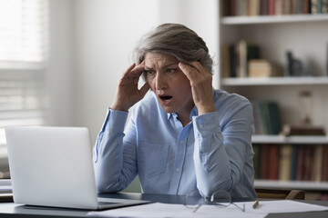 Stressed aged businesswoman staring at laptop hold head feels shocked by bad news, unsaved project...