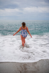 A plump woman in a bathing suit enters the water during the surf. Alone on the beach, Gray sky in the clouds, swimming in winter.