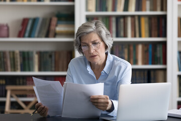 Serious thoughtful middle-aged woman in glasses looks worried read news in formal document sit at workplace desk with wireless computer. Older female review paper letter, learns report feels concerned