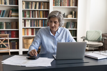 Attractive middle-aged 60s woman accountant calculating sum to pay, seated at workplace desk with laptop and bills. Female managing personal finances, control family incomes, check balance concept