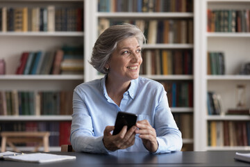Satisfied middle-aged business lady sit at desk staring into distance with smartphone in hands....
