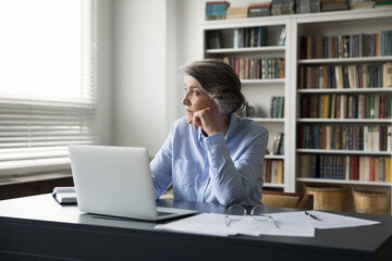 Pensive serious older business lady sit at workplace desk with laptop staring out window looks...