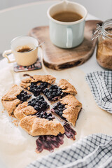Blueberries Galette on table with coffee cup, kettle and sugar jar in background. Simple French recipe, rustic free-form fruit pie, home backing from scratch. Healthy eating lifestyle. Breakfast idea.