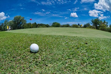 panoramic view of close up white golf ball near green course and one red flag with people in the blue sky