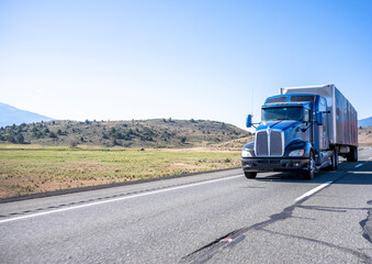 Fototapeta na wymiar Dark blue big rig semi truck with extended cab with roof windows transporting cargo in covered framed semi trailer running on the highway road with hills on the side in California