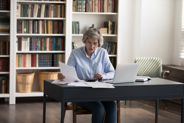 Mature accountant woman sit at workplace desk sorting out bills, making payments, calculates summary for pay through e-bank system on laptop. Checking and management of finances, accounting concept