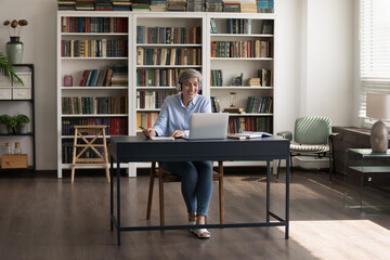 In spacious home office middle-aged woman in headphones writes information in copybook, e-learning...