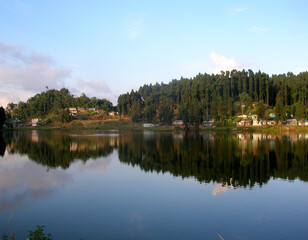 Fototapeta na wymiar The conifer pine trees and houses mirrored at Sumendu Lake look mesmserizing at Mirik in Darjeeling. This is an old artificial lake measuring around 1.25 km long and main attraction of the place.