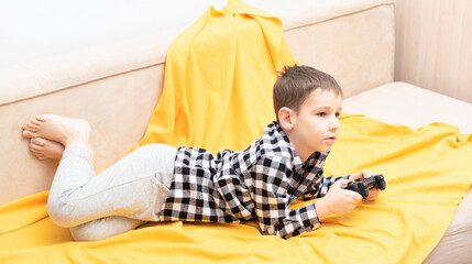 The child boy in checked shirt lying on the couch with black joystick in his hands playing the...