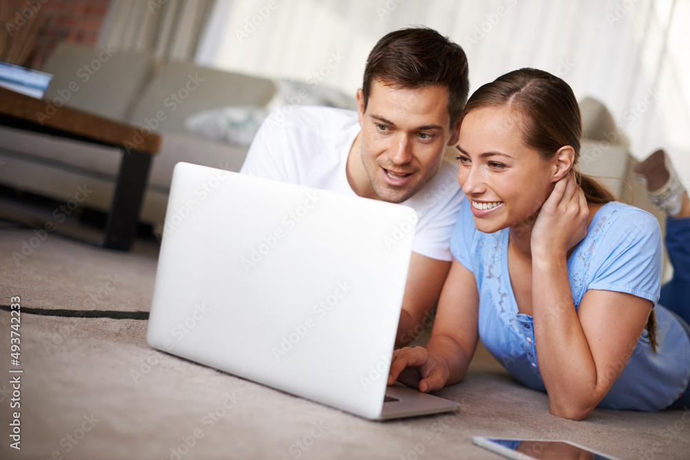 Poster Lets watch that one. A happy young couple lying on their living room floor with a laptop.