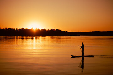 Woman on sup board with reflection on water surface rowing with oar in hands on evening lake with...
