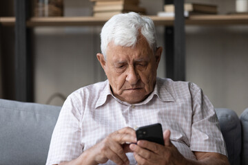 Concerned senior man sit on sofa at home holds smartphone, staring at screen, learn mobile app, experiences troubles with modern tech usage, need help, device malfunction, lack of understating concept
